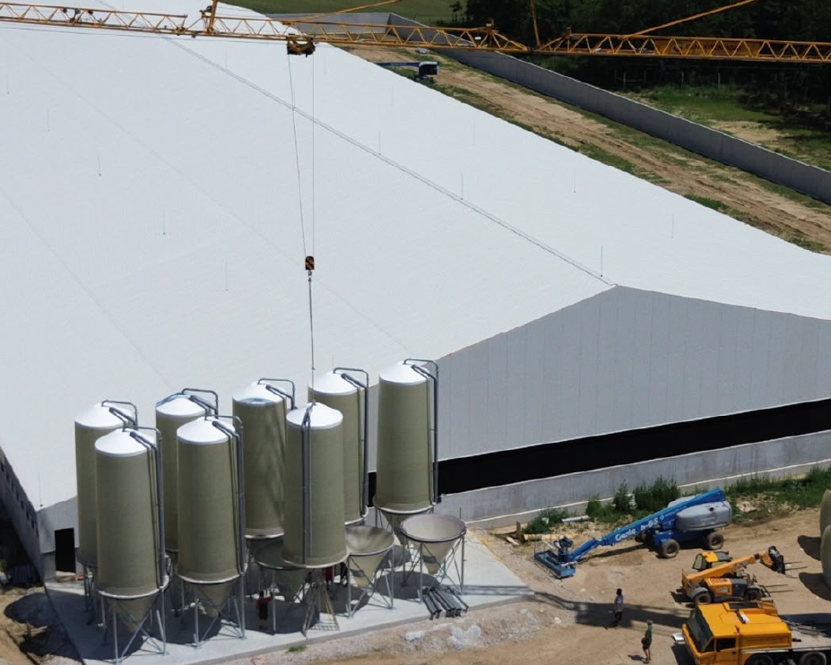 Luchtfoto van plaatsing van hoge, zilverkleurige voersilo's naast een grote witte stal, met behulp van een gele bouwkraan in een rustige, landelijke omgeving.